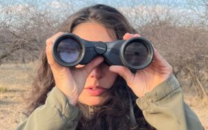A brunette woman facing the camera looking through binoculars surrounded by grassland